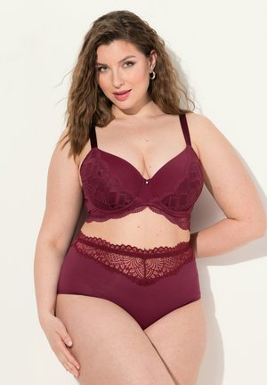 Kobieta z falowanymi włosami, ubrana w bordowy koronkowy biustonosz i majtki z wysokim stanem, stojąca na jasnym tle, pewnie się uśmiechająca.