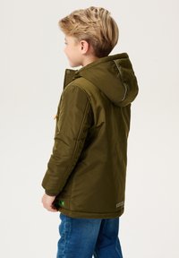 Młody chłopiec z krótkimi blond włosami, ubrany w oliwkową zieloną zimową kurtkę z kapturem i niebieskie dżinsy, zwrócony w lewo na jednolitym tle.
