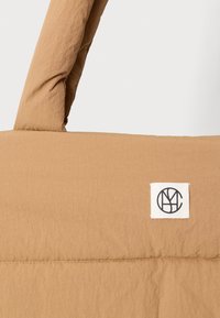 Světle béžová látková taška s polstrovanými rukojeťmi a malou bílou čtvercovou etiketou s černým kruhovým logem na jedné straně.