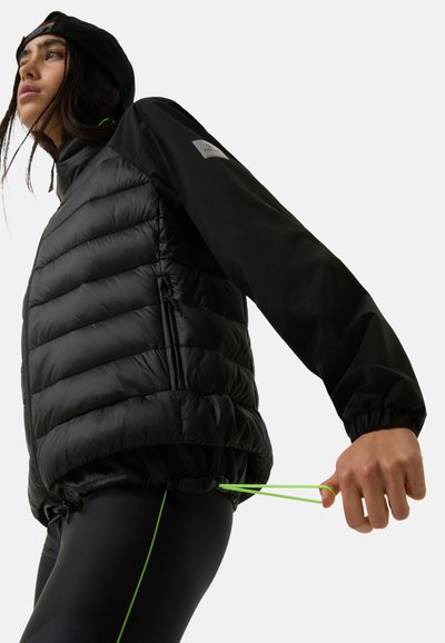 Sort puffjakke med horisontal quiltning, lange ærmer og grønne snore i taljen. Båret over sorte leggings.