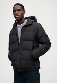 Jachetă puf din negru cu fermoar frontal, glugă și design matlasat. Are buzunare laterale și manșete elastice. Purtați-o peste un pulover gri și blugi.