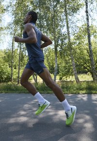Běžecké boty Nike s bílým a neonově zeleným designem, se strukturální podrážkou. Model má na sobě bezsleeve šedý top a šortky.