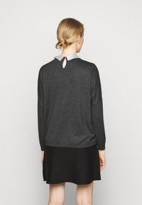 Osoba z blond włosami związanymi w niski kok, ubrana w ciemnoszary długi rękaw, z jasnoszarym kołnierzykiem i czarną spódnicą, odwrócona plecami.