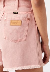 Primer plano de una persona que lleva pantalones cortos de mezclilla rosas con bordes deshilachados y un parche de cuero Wrangler en el bolsillo trasero.