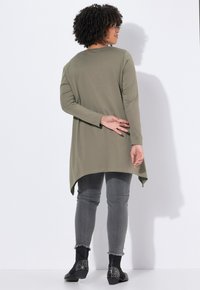 Túnica larga verde olivo con dobladillo curvado, combinada con jeans grises. La modelo lleva botas de tobillo negras con tachuelas, destacando un diseño de ajuste relajado.
