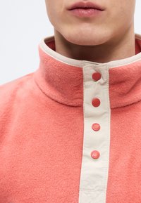 Ζακέτα από φλις με ψηλό γιακά, σε κοραλλί και εκρού χρώμα. Διαθέτει τέσσερα κοραλλί κουμπιά μπροστά και υφασμάτινη υφή.