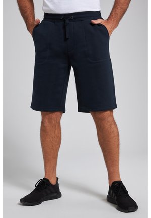 Homme portant un short bleu marine à cordon et des chaussures de sport noires, debout les mains dans les poches.