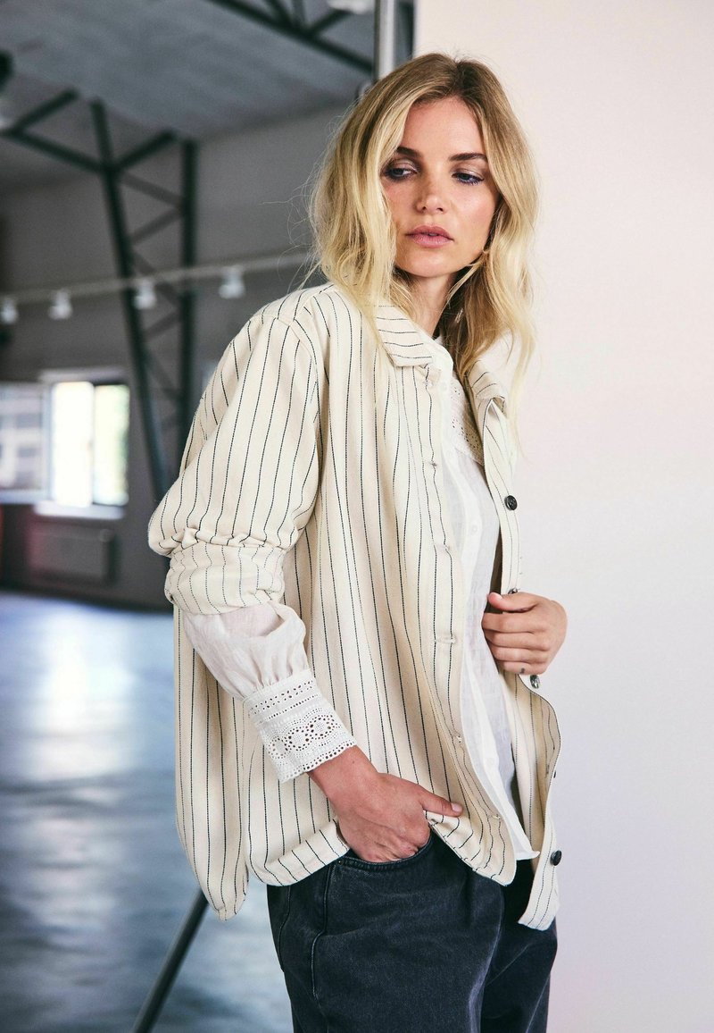 Camicia oversize a righe crema con chiusura a bottoni, dotata di dettagli in pizzo bianco testurizzato sui polsini e una vestibilità rilassata.