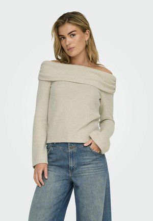 Pull en maille beige clair, dégageant les épaules, avec une texture côtelée, des manches longues et une coupe ajustée, associé à un jean large bleu.