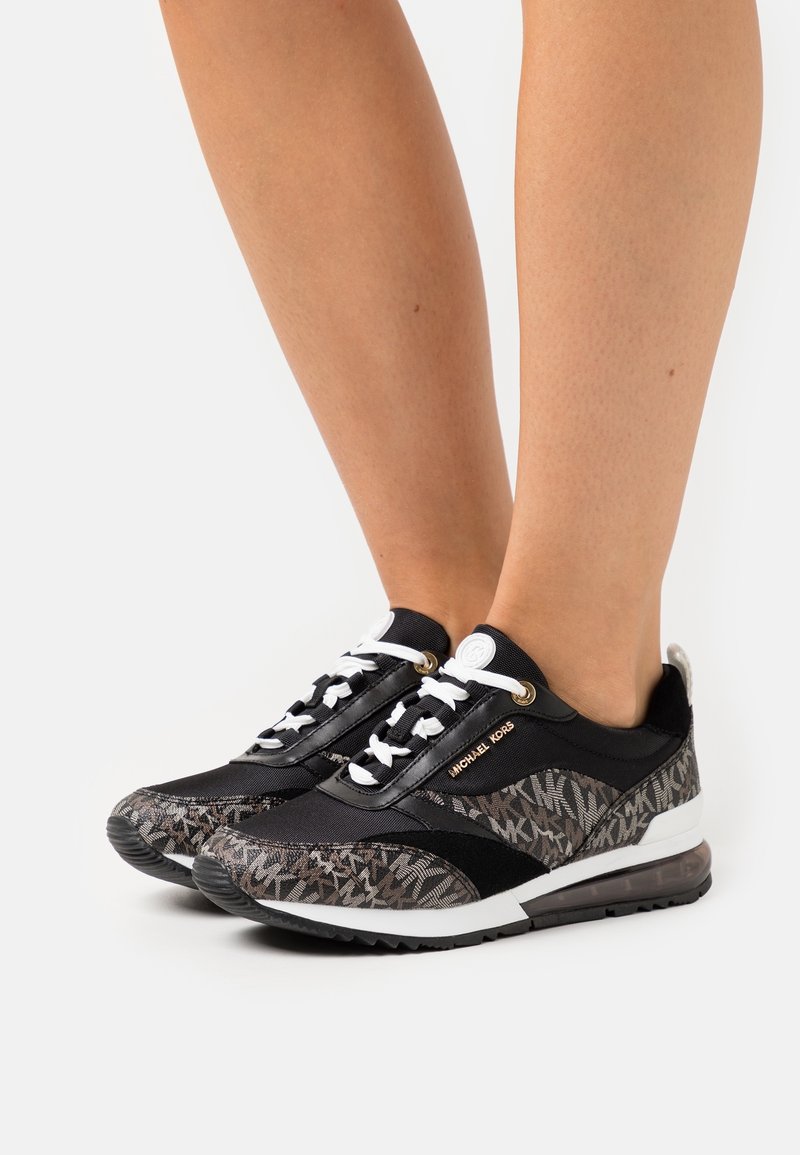 Jambes portant des baskets Michael Kors noires et grises à motifs avec semelles et lacets blancs sur fond blanc.