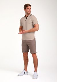 Camisa polo beige con ribetes negros, de mangas cortas y cuello con cremallera, combinada con pantalones cortos marrones con puños y zapatillas blancas. Textura de tela suave.