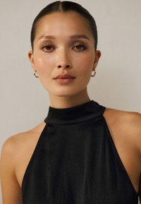 Schwarzes ärmelloses Neckholder-Top aus strukturiertem Stoff mit hohem Kragen und glatten Nahtdetails. Goldene Creolen runden den Look ab.