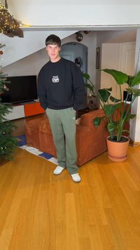 Schwarzer Pullover mit Logo, olivgrüne Hose und weiße Turnschuhe. Innenaufnahme mit braunem, strukturiertem Sofa, grüner Pflanze und Holzboden.