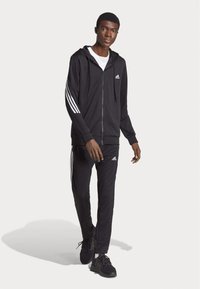Haut à zip noir et pantalon assorti en tissu lisse, avec des accents blancs à trois bandes sur les manches et les jambes. Logo Adidas visible.