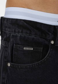 Sorte denim jeans med et lille "Pezador"-mærke, sølv hardware og en hvid talje med grå striber synlige øverst.