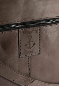 Bruine leren tas met een gestructureerd oppervlak, een ritssluiting en een reliëflabel met een ankerontwerp en het woord "HARBOUR".