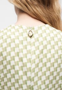 Ξανθά μαλλιά πέφτουν πάνω σε ένα πράσινο και λευκό πουκάμισο με μικρό χρυσό φερμουάρ στην πίσω πλευρά του λαιμού.