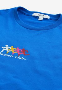 Sininen puuvillapaita, jossa on pyöreä kaula-aukko. Paidassa on värikkäitä juoksevia hahmoja vaaleanpunaisena, keltana ja valkoisena, sekä teksti "Runners Club=" alareunassa.
