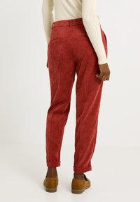 Persona vestida con pantalones de pana color óxido, suéter de canalé beige y mocasines de ante marrón, de pie contra un fondo claro y liso.