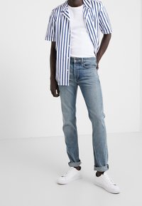 Camisa blanca de manga corta con rayas verticales azules, combinada con jeans de color azul claro y zapatillas blancas. Atuendo casual con un ajuste relajado.