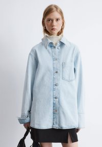 Light blue denim oversized jacket with a pointed collar, button-down front, single chest pocket, and rolled cuff sleeves.
