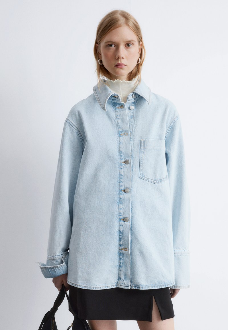 Light blue denim oversized jacket with a pointed collar, button-down front, single chest pocket, and rolled cuff sleeves.