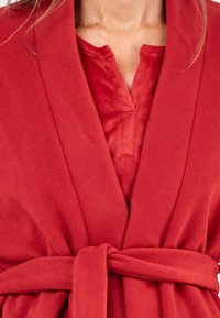 Bata de felpa roja con cuello chal, cinturón en la cintura y abertura frontal. Textura suave con un acabado liso. Presenta un botón en el escote.