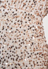 Vitt tyg med en texturerad yta, prytt med växlande guld- och svarta paljetter i olika storlekar, vilket skapar en skimrande effekt.