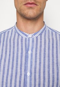 Button-up shirt featuring vertical blue and white stripes, a band collar, and two white buttons at the neck. Cotton or linen texture.