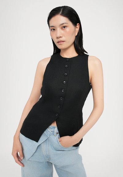 Sleeveless black linen top with a high neckline, featuring a front button closure and a split design at the hem. Worn with light blue jeans.