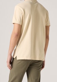 Polo beige, à manches courtes, en coton doux avec des fentes sur le bas. Assorti à un pantalon vert olive, texture lisse visible.