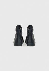 Une paire de bottines en cuir noir avec fermeture éclair à l'arrière et languette, présentée de l'arrière sur un fond gris clair uni.