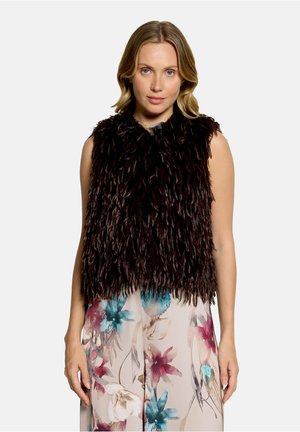 Vrouw met blond haar draagt een mouwloze donkerbruine, pluizige bodywarmer over een beige jurk met grote blauwe, roze en witte bloemenprints.