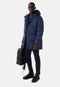 Navy blue puffer coat with a hood, zippered front, and side pockets. Worn with dark pants and black backpack, complemented by black sneakers.