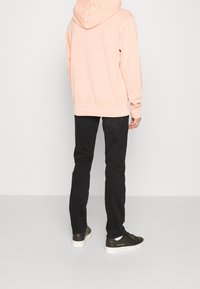 Osoba v světle růžové mikině, černých úzkých kalhotách a černých teniskách stojí k nám zády proti jednobarevnému bílému pozadí.