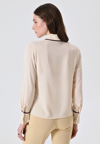 Donna con lunghi capelli castani, che indossa una blusa di seta crema con rifiniture scure e pantaloni beige, rivolta verso sinistra su uno sfondo semplice.