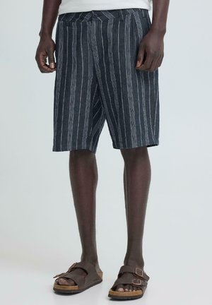 Mann trägt dunkelgraue und weiße vertikal gestreifte Shorts und braune Sandalen mit doppeltem Riemen, steht vor einem einfarbigen Hintergrund.