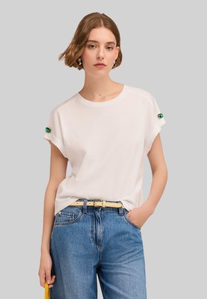 Tricou alb cu mânecă scurtă și manșete răsucite, cu decorațiuni verzi, asortat cu blugi albaștri cu talie înaltă și o geantă galbenă.