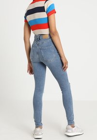 Henkilö, joka on pukeutunut korkeavyötäröisiin vaaleansinisiin skinny-farkkuihin, lyhythihaiseen raidalliseen paitaan, jossa on punaista, sinistä ja mustaa, sekä valkoisiin tennareihin, seisoo yksinkertaista taustaa vasten.