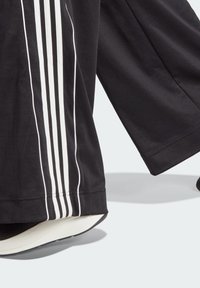 Black athletic pants featuring three white stripes along the side, with a soft fabric texture and a loose fit, paired with a black sneaker.