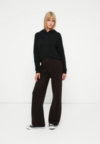 Femme aux cheveux blonds portant un sweat à capuche noir, un pantalon large marron foncé et des baskets noir et blanc, se tenant devant un fond blanc uni.