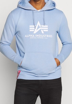 Mężczyzna w jasnoniebieskiej bluzie z kapturem Alpha Industries z logo i napisem na piersi, ręce w przedniej kieszeni, w połączeniu z czarnymi spodniami.