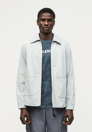 Mann iført en lys grå jakke med glidelås over en mørkeblå grafisk t-skjorte og grå cargobukser, stående mot en ensfarget bakgrunn.