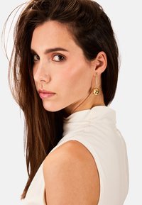 Pendientes de gota en tono dorado que presentan un acento esférico y un poste delgado y alargado. La modelo tiene el cabello largo y lacio, y lleva una blusa de cuello alto.