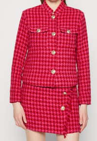 Giacca a motivi houndstooth rossi con bottoni dorati, caratterizzata da due tasche frontali e una gonna coordinata con frange e tre bottoni di lato.