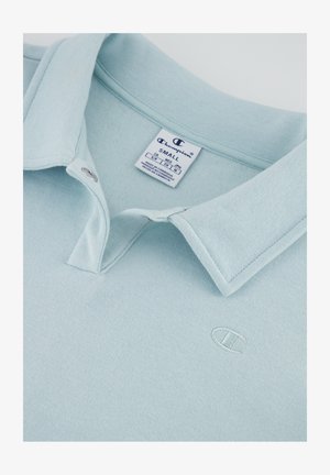 Polo shirt azzurro chiaro con una texture morbida, dotato di colletto con bottoni e un piccolo logo ricamato sul petto. L'etichetta indica la taglia S.
