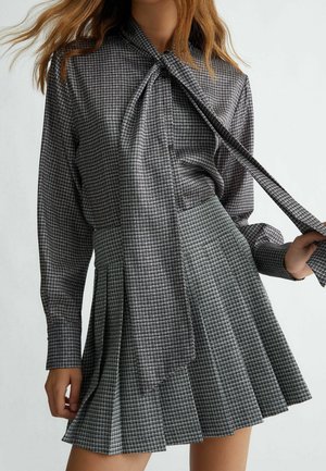 Kobieta ubrana w dopasowaną szarą kratkowaną bluzkę z długim rękawem i wiązaniem przy szyi oraz plisowaną spódnicę, trzymająca wiązanie jedną ręką.