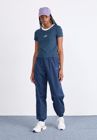 Ναυτικό μπλουζάκι με λευκές λεπτομέρειες και κείμενο "Nike", συνδυασμένο με χαλαρούς ναυτικούς παντελόνες. Το μοντέλο φοράει ένα ανοιχτό μωβ καπέλο και λευκά αθλητικά παπούτσια.