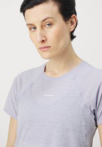Violet sportinė marškinė su trumpomis rankovėmis, pasižyminčios lygiu paviršiumi ir maža balta logo krūtinėje. Paprastas, prigludęs dizainas.
