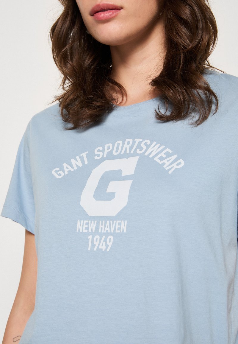 Ljusblå bomullströja med korta ärmar och rund halsringning, prydd med ett vitt grafiskt tryck: "GANT SPORTSWEAR," ett stort "G," och "NEW HAVEN 1949."